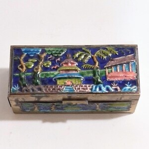 May include: A rectangular metal box with a hinged lid. The lid is decorated with a colorful enamel scene featuring a pagoda, trees, and a building against a blue background. The box has a silver-colored metal frame.
