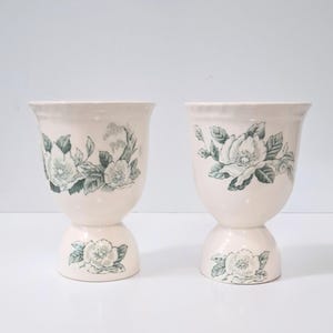 May include: Two off-white ceramic vases with a floral design. The vases have a pedestal base and a flared top. The floral design features white flowers and green leaves. The vases are displayed on a white surface.