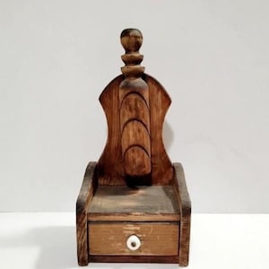 May include: A small, wooden decorative box with a drawer and a tall, ornate back. The box is stained a warm brown color, with a small white knob on the drawer. The back features a carved design.