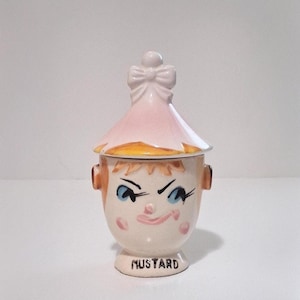 May include: A vintage ceramic mustard jar with a whimsical face design. The jar features a pink and white hat-shaped lid with a bow, and a smiling face with blue eyes and rosy cheeks. The word "MUSTARD" is printed on the base.