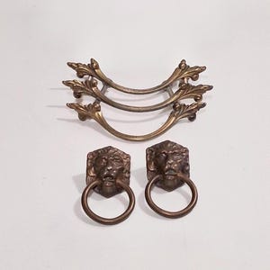 May include: Three curved brass drawer pulls with ornate leaf-like ends stacked vertically, accompanied by two brass lion head drawer pulls featuring circular rings hanging from the mouths of the lions, all with a metallic antique finish.