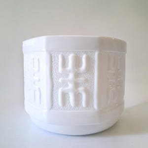 May include: A white cylindrical planter featuring a repeating embossed geometric pattern on its exterior. The surface texture alternates between smooth vertical panels and textured panels with raised, stylized symbols in a bright white finish.