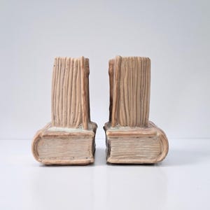 May include: A pair of bookends, each designed to resemble a stack of books. The bookends are a light beige color with a textured, vertical design on the spine and a rounded edge on the bottom, giving them a classic, vintage look.