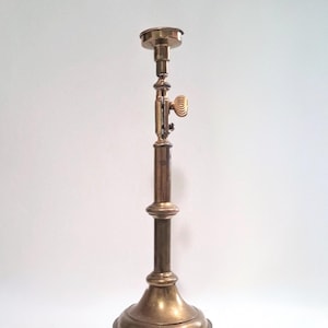 May include: A vintage brass candlestick with an adjustable height mechanism. The candlestick has a round base, a long central stem, and a small cup at the top for holding a candle. The adjustable section features a small, decorative handle.