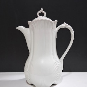 May include: A white porcelain coffee pot featuring a vertical ribbed design, a curved handle, a pointed spout, and a lid topped with a decorative finial. The body of the pot has a scalloped base and a smooth, glossy finish throughout the entire piece.