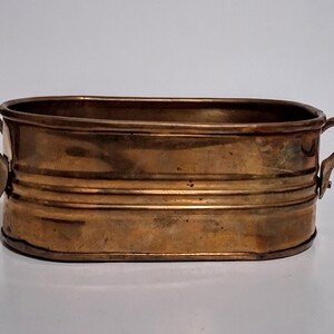 May include: An oval-shaped brass planter with a polished metallic finish and a warm golden-brown hue. The vessel features two curved side handles attached with rivets and horizontal ribbed detailing along the exterior walls of the container.