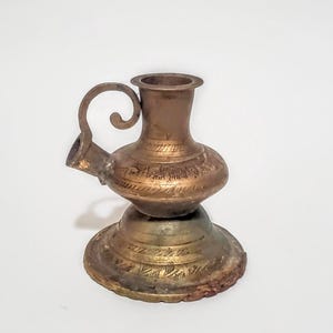 May include: A metallic candle holder with a brass-toned finish, featuring a wide circular base, a bulbous central body, and a curved handle. The surface is decorated with etched horizontal lines and patterns around the base and midsection.
