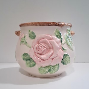 May include: A ceramic planter with a light pink base, adorned with a large, raised pink rose and green leaves. The rim has a brown, woven texture, and it features two small side handles. A decorative floral container.