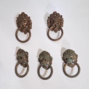 May include: A set of five bronze-toned metal drawer pulls, each featuring a detailed lion head design with a circular ring handle attached. The hardware displays a weathered finish with varying levels of oxidation and patina across the surfaces.