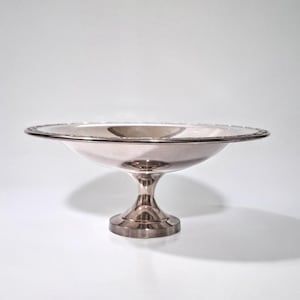 May include: A silver-colored, pedestal serving bowl with a wide, shallow dish and a decorative rim. The bowl sits on a short, flared pedestal base. The bowl is likely made of metal and reflects the light.