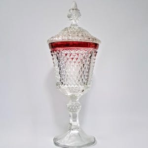 May include: A clear glass pedestal candy dish with a diamond pattern and a red rim. The dish has a lid with a pointed finial. The base is a wide, clear, footed design.