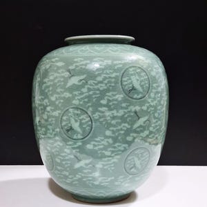 May include: A celadon-colored ceramic vase with a round body and a slightly flared rim. The vase features a design of cranes and clouds. The cranes are depicted in flight, and the clouds are rendered in a stylized manner. The vase is approximately 12 inches tall.