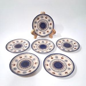May include: A set of six ceramic plates with a cream-colored background, blue and brown decorative patterns, and a dark blue center. One plate is displayed on a gold-colored plate stand. The plates have a circular design with heart and dot motifs.