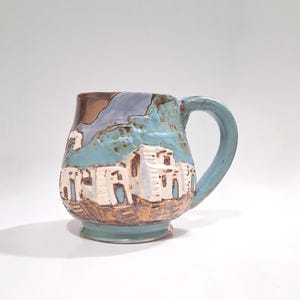 May include: A ceramic mug with a hand-painted village scene. The mug features a turquoise handle and base, with a textured design of white buildings, brown earth, and blue sky. The mug is approximately 4 inches tall.