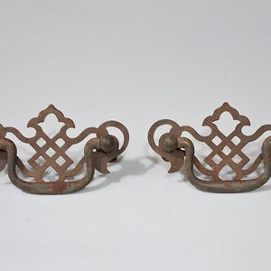 May include: Two antique, ornate drawer pulls with a distressed, rusty finish. Each pull features a decorative openwork design with a central diamond pattern and curved handles. Ideal for vintage furniture restoration projects.