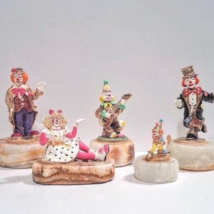 May include: A collection of five clown figurines, each mounted on a polished, banded stone base. The clowns feature colorful outfits, including polka-dot dresses, formal suits, and hats, with painted facial details and various poses, such as playing a guitar.