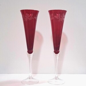 May include: A pair of tall champagne flutes featuring deep red glass bowls with a delicate etched holly leaf pattern near the rim. Each flute has a slender, clear glass stem and a circular clear glass base, standing upright on a flat surface.