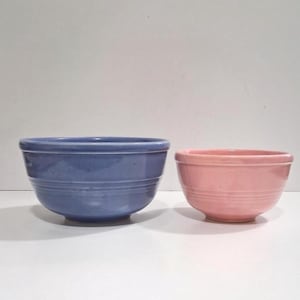 May include: Two ceramic bowls, one periwinkle blue and one light pink, sit side-by-side. The bowls have a rounded shape with a slightly flared rim and horizontal grooves around the body. The bowls are on a white surface.