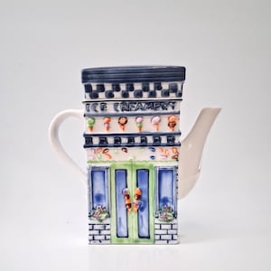 May include: A ceramic teapot shaped like an ice cream shop, featuring a white handle and spout. The building design includes the text "ICE CREAMERY", with colorful ice cream cone decorations and a brick facade. The color scheme includes blue, green, and white.
