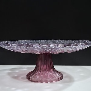Pu&ograve; includere: Un alzatina per torte in vetro trasparente color lavanda, con bordo smerlato e base a coste. L'alzatina &egrave; rialzata e presenta un motivo decorativo sul bordo. Lo sfondo &egrave; bianco e nero.