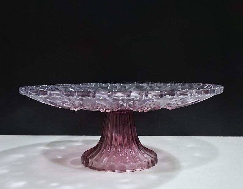 Pu&ograve; includere: Un alzatina per torte in vetro trasparente color lavanda, con bordo smerlato e base a coste. L'alzatina &egrave; rialzata e presenta un motivo decorativo sul bordo. Lo sfondo &egrave; bianco e nero.
