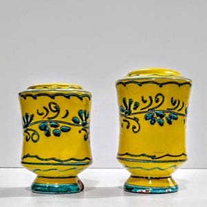 May include: Two yellow ceramic vases with a floral design in teal and black. The vases have a rounded shape with a flared base and a lid. The vases are decorated with teal and black floral patterns.