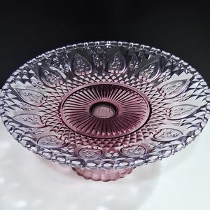 Pu&ograve; includere: Un'alzatina per torte vintage in vetro trasparente con una tonalit&agrave; rosa. L'alzatina presenta un design dettagliato in rilievo con un centro circolare e un bordo smerlato. La base &egrave; di colore rosa abbinato e il design generale &egrave; ornato.