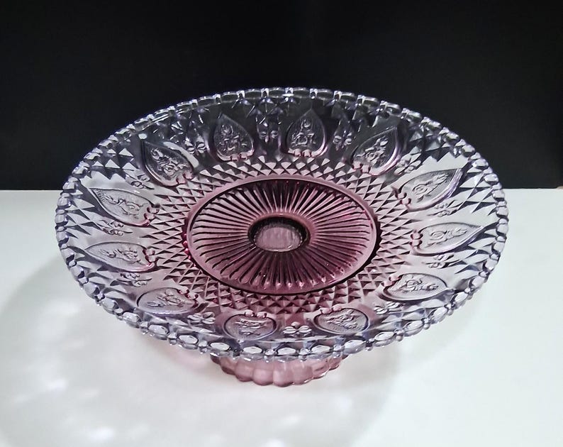 Pu&ograve; includere: Un'alzatina per torte vintage in vetro trasparente con una tonalit&agrave; rosa. L'alzatina presenta un design dettagliato in rilievo con un centro circolare e un bordo smerlato. La base &egrave; di colore rosa abbinato e il design generale &egrave; ornato.