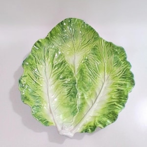 May include: A ceramic serving plate shaped like a large lettuce leaf. The dish features realistic textures and a gradient of green and white colors, with prominent raised veins and ruffled edges that mimic the appearance of fresh garden greens.