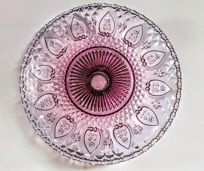 Pu&ograve; includere: Un piatto da portata rotondo in vetro vintage con un design decorativo. Il piatto presenta un cerchio centrale magenta scuro circondato da un'area rosa pi&ugrave; chiara con motivi a forma di foglia. Il bordo ha un bordo strutturato e perlato. Per servire cibo.