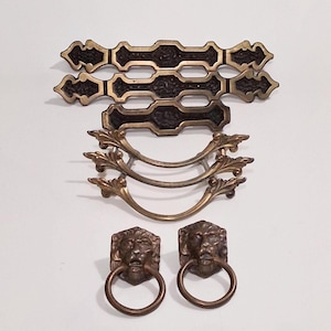 May include: An assortment of antique bronze-toned furniture hardware. The collection includes two long, ornate pulls, three curved pulls with leaf accents, and two lion head ring pulls. The hardware is arranged on a white background.