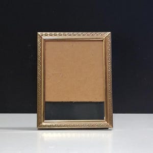 May include: A rectangular gold-colored picture frame with a decorative border. The frame has a brown backing and a black section at the bottom. The frame is standing on a white surface against a black background.