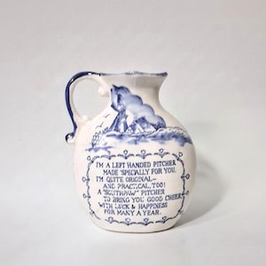 May include: A cream-colored ceramic pitcher with a blue handle and blue nautical illustrations. The text reads: I'M A LEFT HANDED PITCHER MADE 'SPECIALLY FOR YOU. I'M QUITE ORIGINAL- AND PRACTICAL, TOO! A "SOUTHPAW" PITCHER TO BRING YOU GOOD CHEER WITH LUCK & HAPPINESS FOR MANY A YEAR.