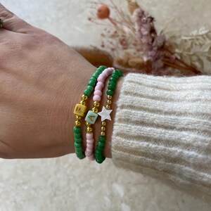 May include: Three beaded bracelets on a wrist. The bracelets are green, pink, and gold. The gold bracelet has a star charm and a letter 'A' charm. The green bracelet has a letter 'E' charm.
