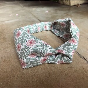 May include: A white headband with a floral pattern featuring pink flowers and green leaves. The headband is made of fabric and has a knotted design.