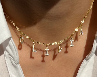May include: A gold chain necklace with the name "Olivia" spelled out in individual charms. Each charm is encrusted with small, clear rhinestones.