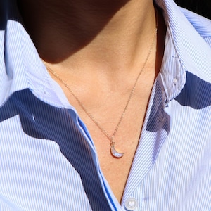 Puede incluir: Un collar de cadena dorada con un colgante de media luna hecho de &oacute;palo blanco iridiscente. El collar lo lleva una persona que lleva una camisa de rayas azul claro.