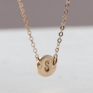 Może przedstawiać: Złoty łańcuszek z małym, okrągłym wisiorkiem z wygrawerowaną literą "S".