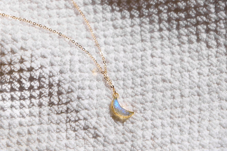 Puede incluir: Un collar de cadena dorada con un colgante de media luna hecho de piedra de luna iridiscente.