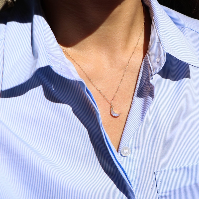 Puede incluir: Un collar de cadena dorada con un colgante de media luna hecho de &oacute;palo blanco iridiscente. El collar lo lleva una persona que lleva una camisa de rayas azul claro.