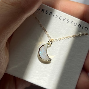 Puede incluir: Un collar de cadena dorada con un colgante de media luna hecho de piedra lunar blanca iridiscente. El colgante est&aacute; engastado en oro y tiene un dise&ntilde;o delicado y femenino. El collar se muestra en una tarjeta blanca con el texto "UREPIECESTUDIO".