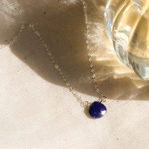 May include: A delicate silver necklace featuring a small, faceted, deep blue gemstone pendant. The pendant is teardrop-shaped and hangs from a fine chain. The necklace is displayed on a neutral-colored fabric, with a glass object in the background.