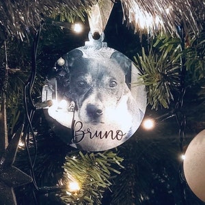 May include: A round, silver Christmas ornament with a black and white photo of a dog. The ornament has the name "Bruno" written on it.