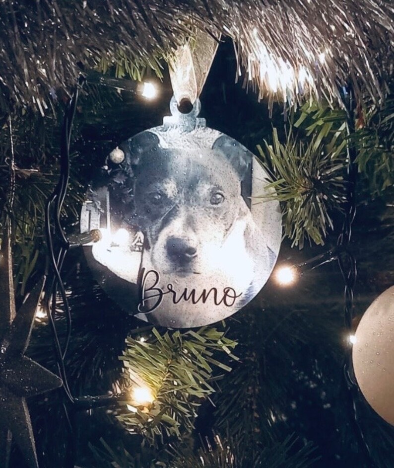 May include: A round, silver Christmas ornament with a black and white photo of a dog. The ornament has the name "Bruno" written on it.