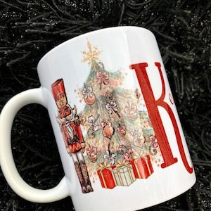 May include: White ceramic mug with a Christmas tree design featuring a nutcracker, presents, and ornaments. The mug has a red letter "K" on the side.