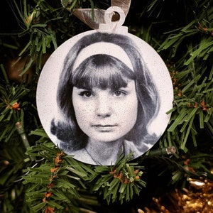 May include: A white round Christmas ornament with a black and white photo of a woman with a headband and bangs.