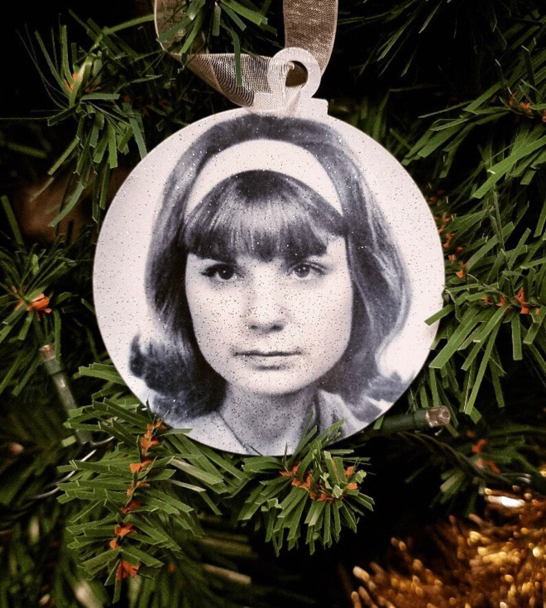 May include: A white round Christmas ornament with a black and white photo of a woman with a headband and bangs.