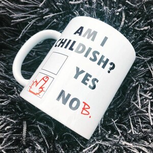 K&ouml;nnte beinhalten: Wei&szlig;e Keramiktasse mit dem Text "Am I Childish?" und einem Kontrollk&auml;stchen mit zwei Optionen: "Yes" und "No B." Die Option "No" ist mit einem roten Mittelfinger angekreuzt.