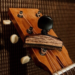 Pode incluir: Um capo de guitarra de madeira com acabamento em madeira marrom escuro e uma botão de ajuste preto. O capo está preso ao braço de uma guitarra acústica com acabamento em madeira natural.
