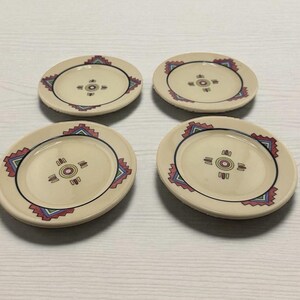 May include: Four small, off-white ceramic plates with a decorative geometric border in red, blue, and green. Each plate features a central design with concentric circles and lines. The plates are arranged on a light-colored surface.